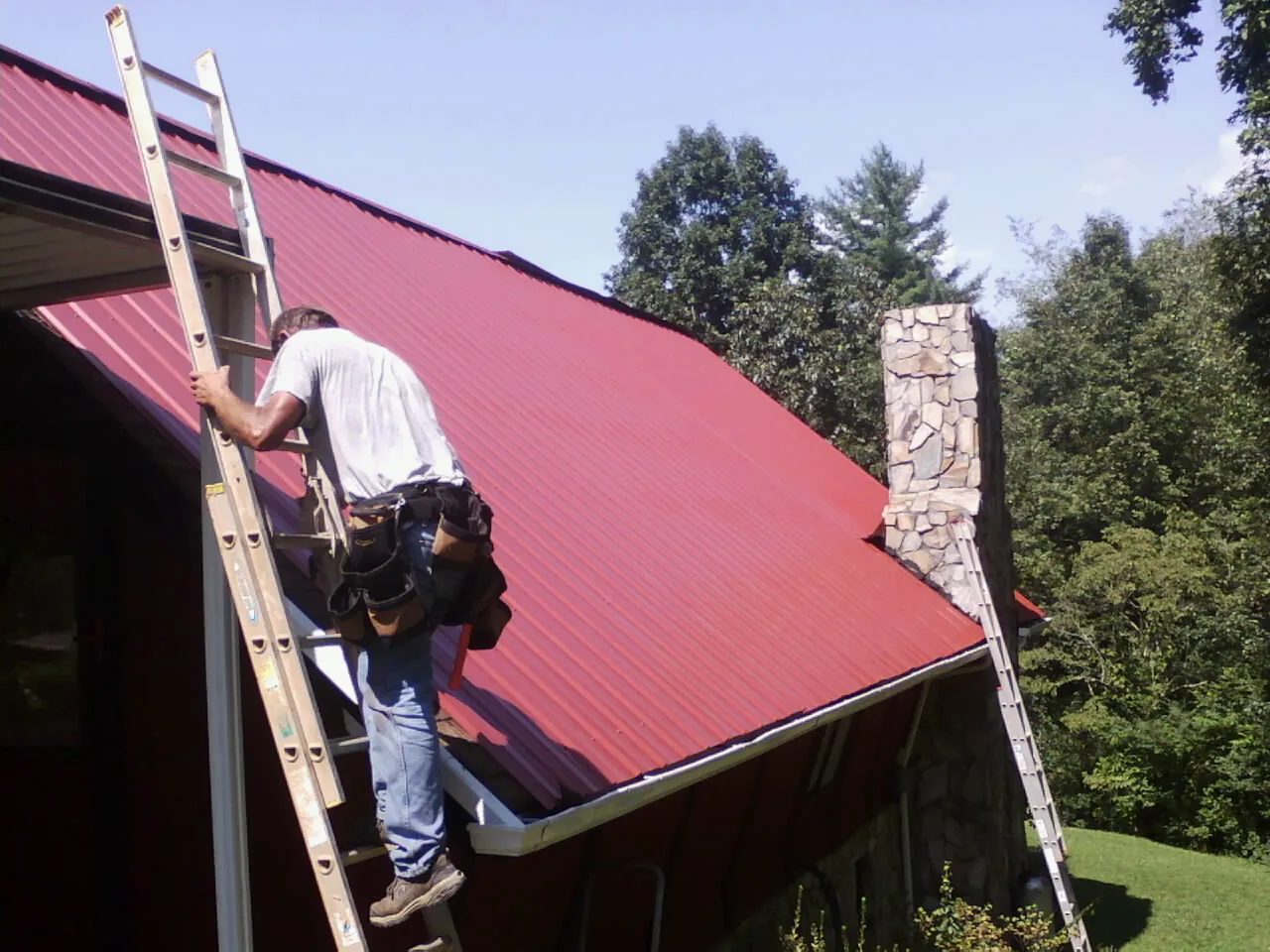 Roofer on a ladder beside a red metal roof and stone chimney — Soffit & Fascia Repair by Roofing Brothers in Milnor